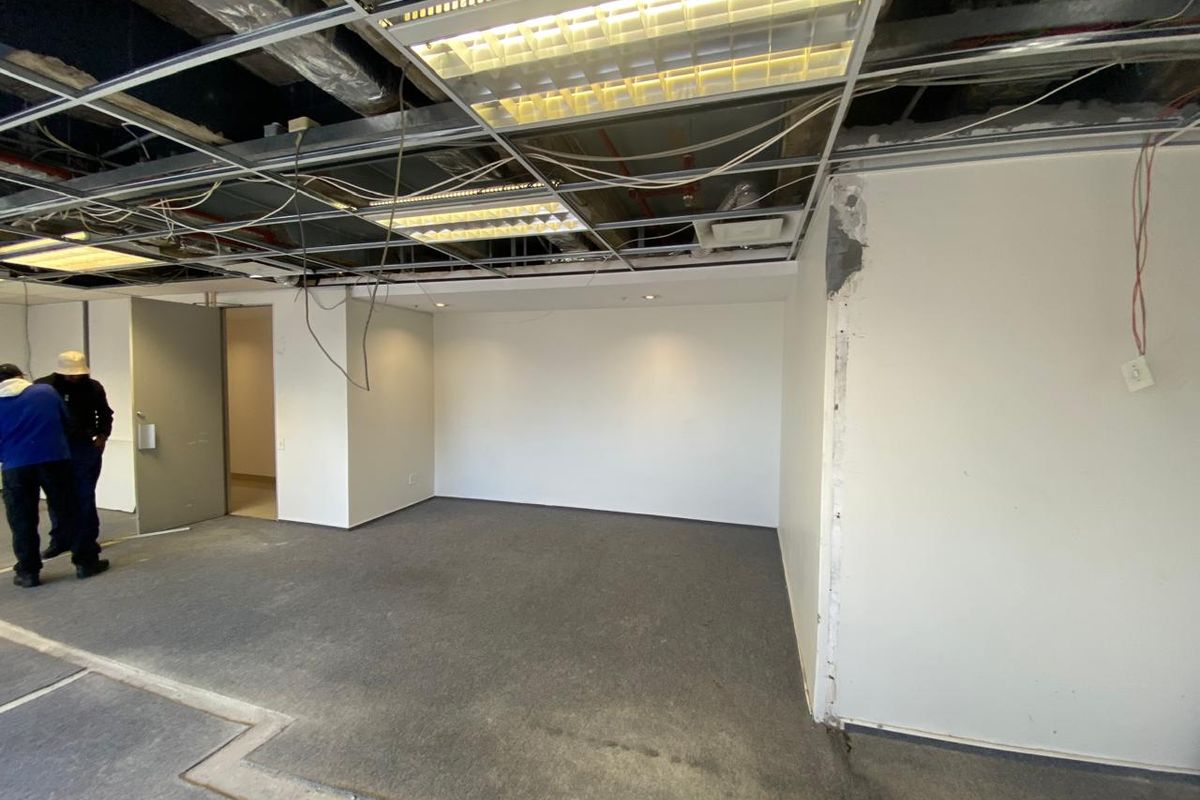 An empty office space undergoing renovation, featuring a partially exposed suspended ceiling, carpeted flooring, and unfinished wall surfaces.