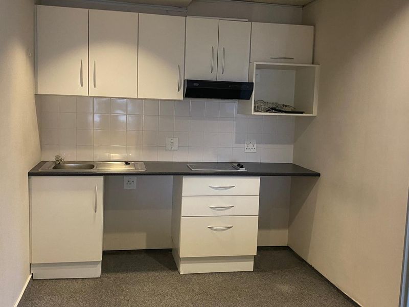 A compact kitchenette featuring white built-in cabinetry, a dark laminate countertop, stainless steel sink, and an integrated electric hob with an extractor fan.