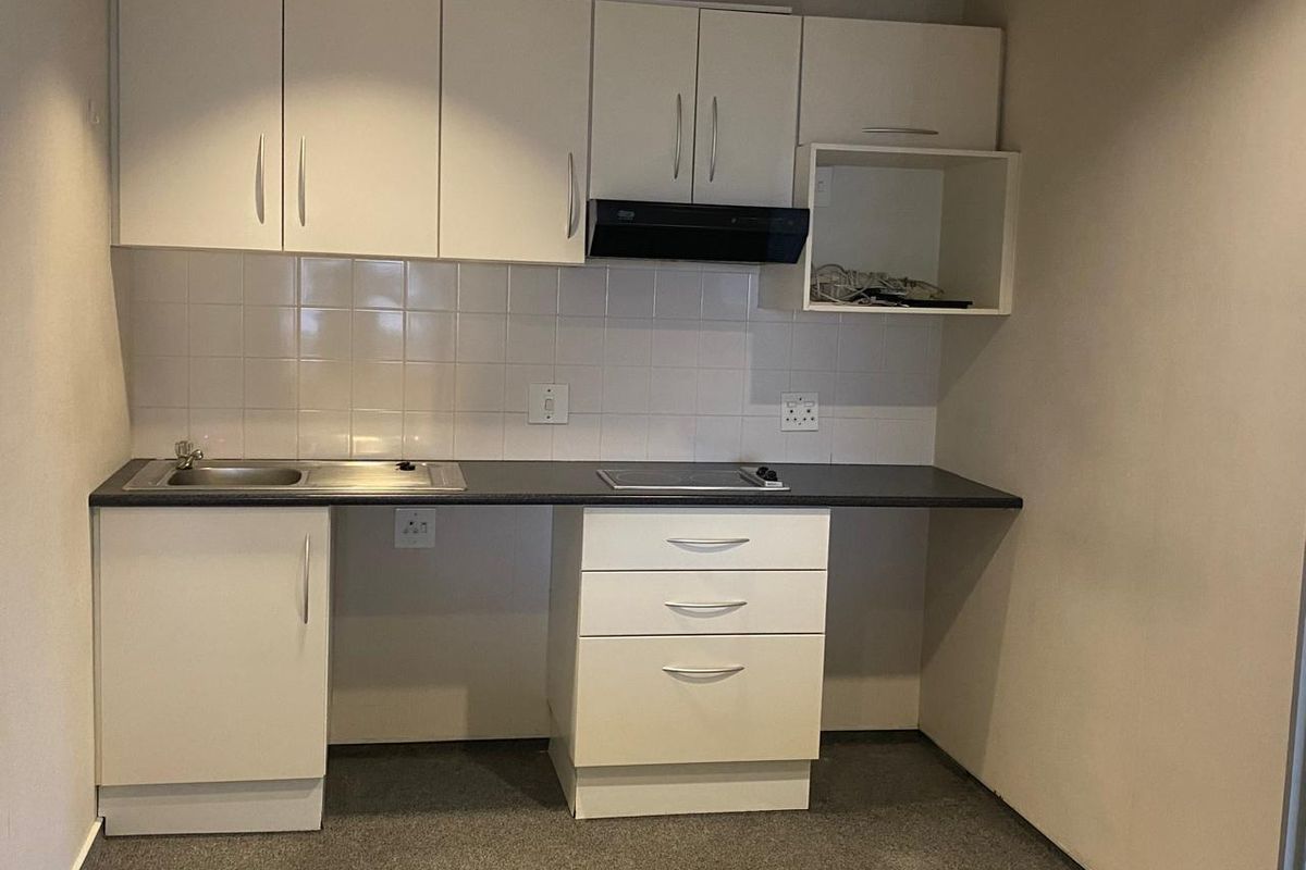 A compact kitchenette featuring white built-in cabinetry, a dark laminate countertop, stainless steel sink, and an integrated electric hob with an extractor fan.