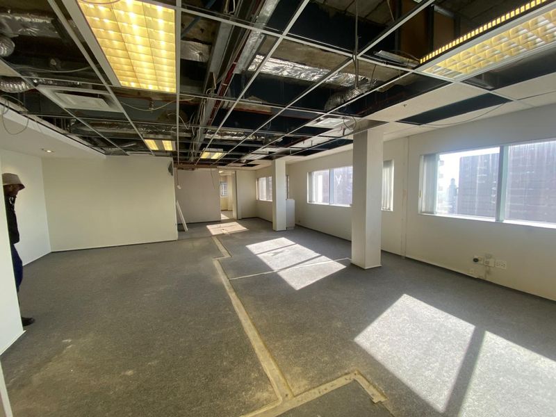 An empty office space featuring large windows, carpeted flooring, and an unfinished suspended ceiling grid with exposed infrastructure.