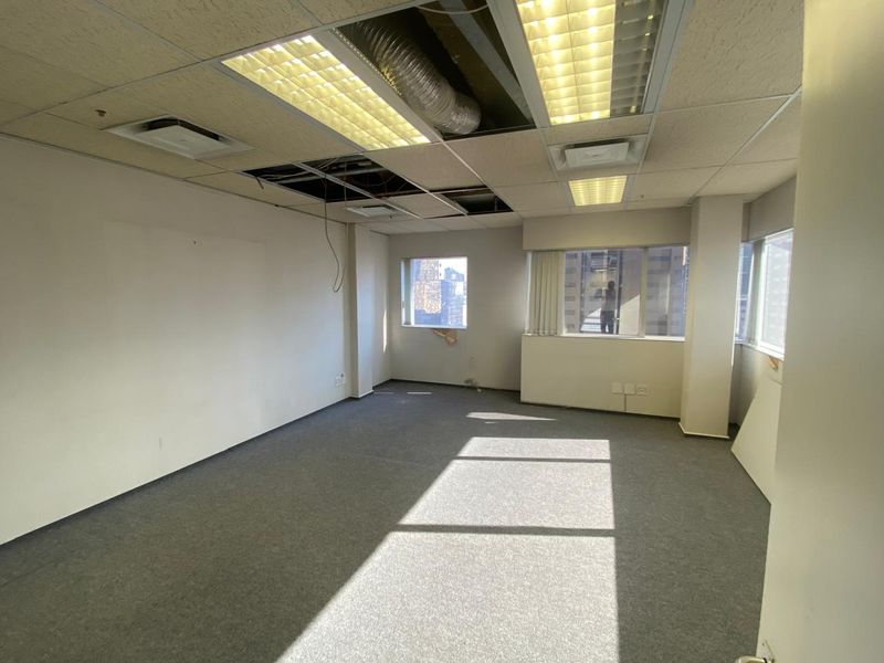 An empty office space featuring large windows, carpeted flooring, and a suspended ceiling with visible maintenance requirements.