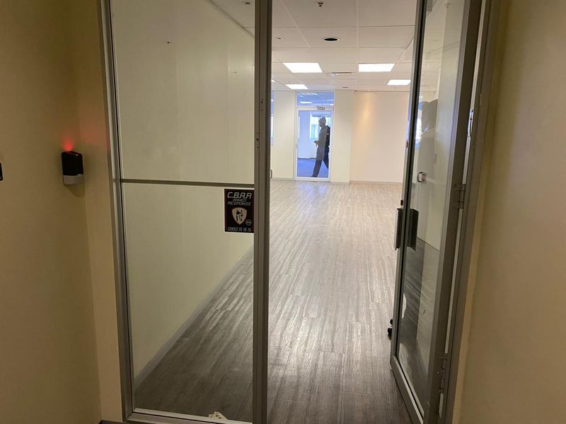 An open-plan office space featuring wood-look vinyl flooring, a suspended ceiling with recessed lighting, and a glass entrance door with access control.