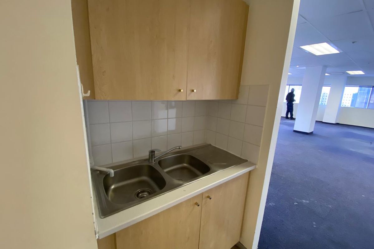 A compact kitchenette area featuring a stainless steel double sink, laminate cabinetry, and tiled splashback, adjacent to an open-plan office space.