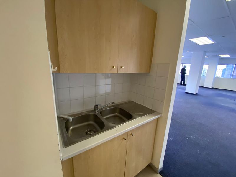 A compact kitchenette area featuring a stainless steel double sink, laminate cabinetry, and tiled splashback, adjacent to an open-plan office space.