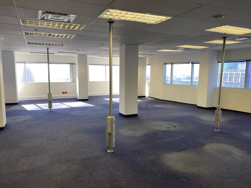 An open-plan office space featuring a suspended ceiling, recessed lighting, and multiple power poles, with large windows providing natural light.