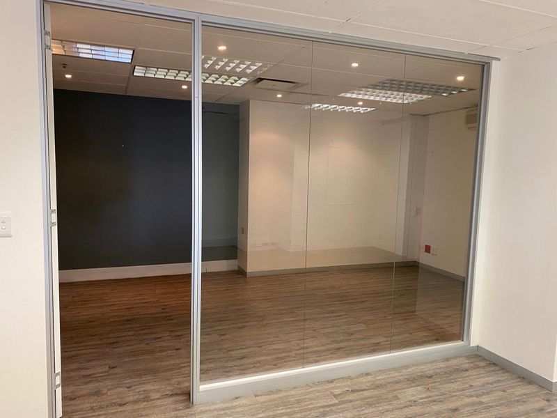 A modern, empty office space featuring a glass partition wall, recessed lighting, and wood-look laminate flooring, suitable for a professional workspace.