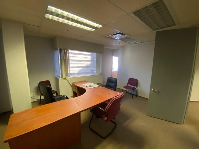 A functional office space featuring carpeted flooring, suspended ceiling with recessed lighting, and large windows with blinds, suitable for a professional work environment.