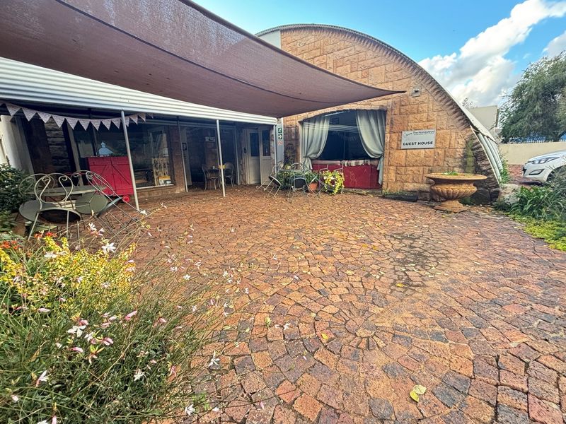 A paved outdoor patio area featuring a shade sail, stone-clad building facade, and established garden surroundings at the guest house entrance.