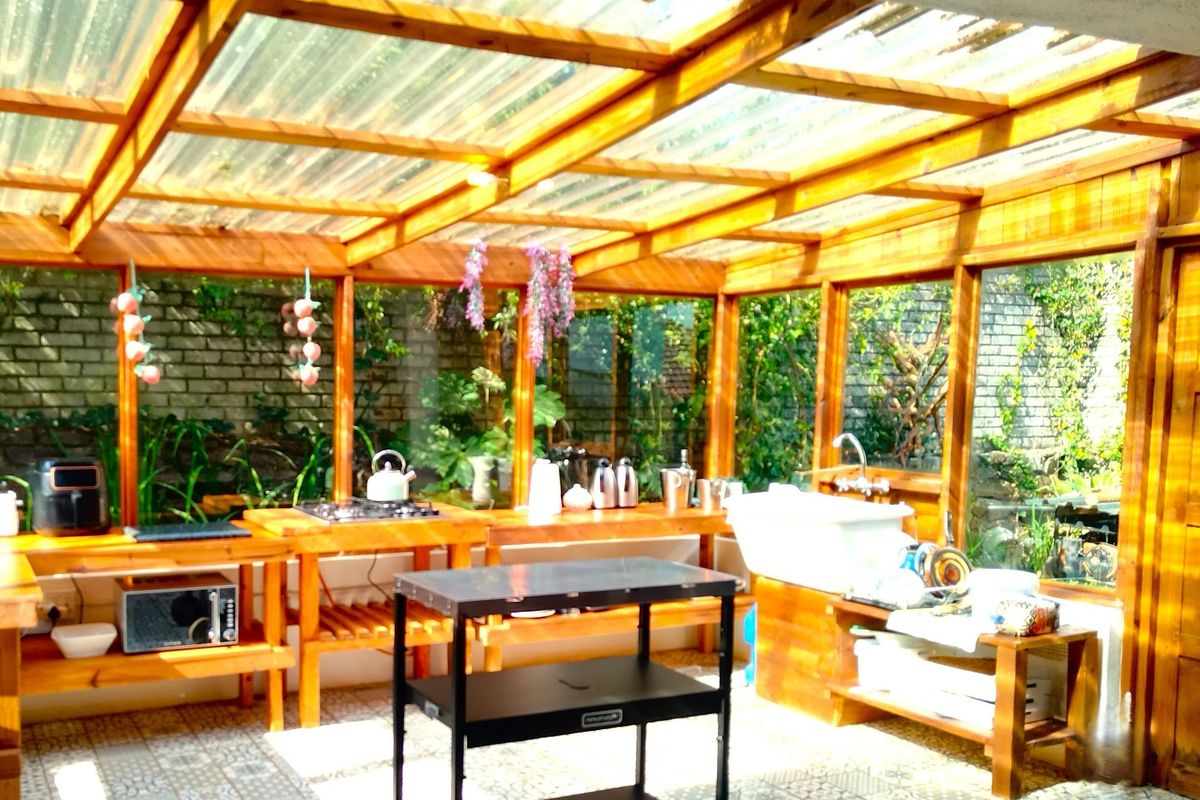 A rustic, light-filled outdoor kitchen featuring a timber pergola roof, gas hob, and farmhouse sink, set within a glass-enclosed garden structure.