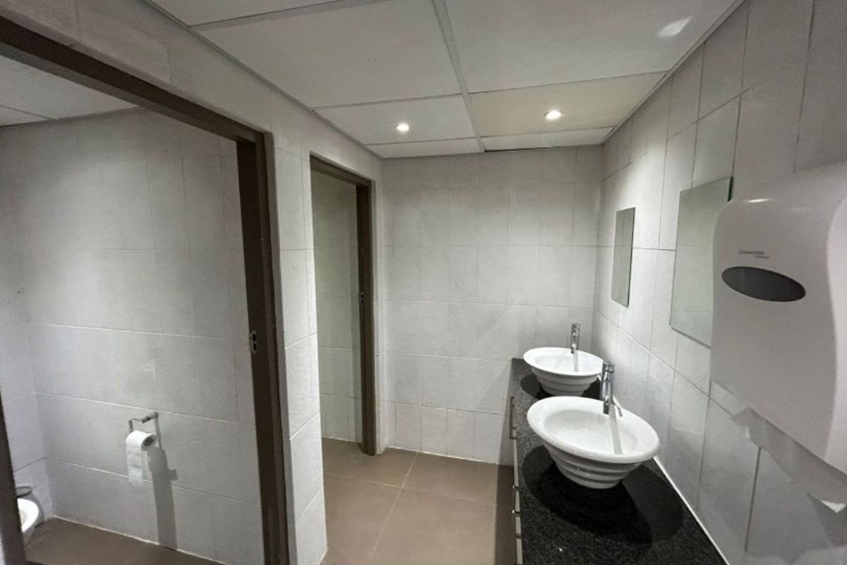 A functional office restroom featuring dual vessel sinks on a granite vanity, tiled walls, and a suspended ceiling with recessed lighting.