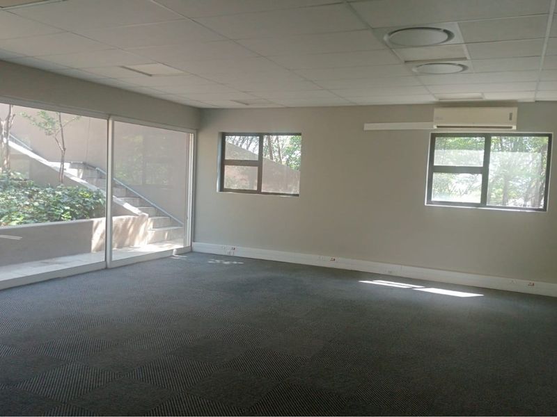 Spacious, empty office suite featuring large windows, carpeted flooring, suspended ceiling tiles, and a wall-mounted air conditioning unit.