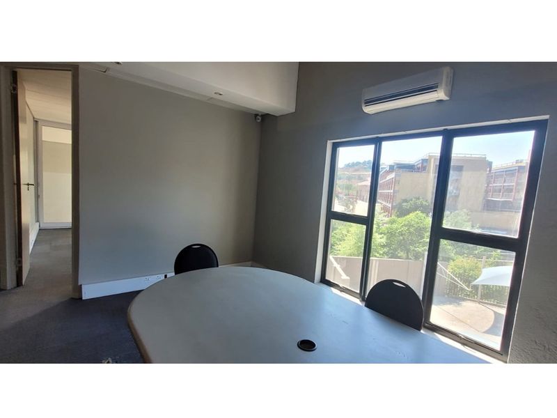 A functional office space featuring large windows with natural light, a wall-mounted air conditioning unit, and carpeted flooring.