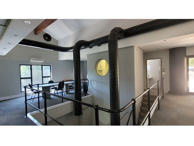 An open-plan office space featuring industrial-style exposed ducting, vaulted ceilings with timber beams, carpeted flooring, and a mezzanine-level layout with metal railings.