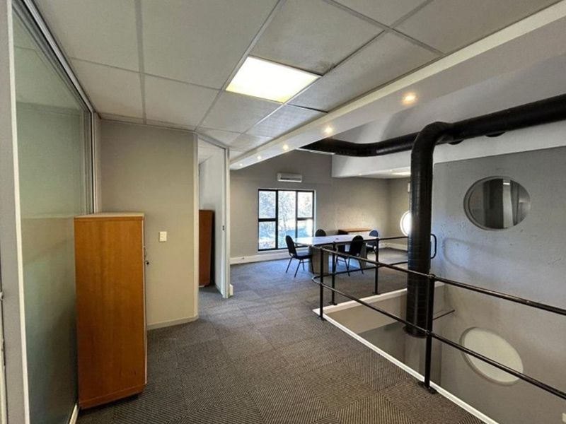 An open-plan office mezzanine area featuring industrial-style exposed ducting, carpeted flooring, and a glass-partitioned office space overlooking the lower level.