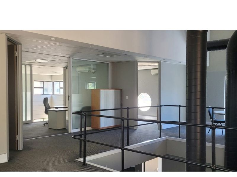 Open-plan office space featuring industrial-style exposed ducting, glass-partitioned private offices, and carpeted flooring with a mezzanine-style layout.