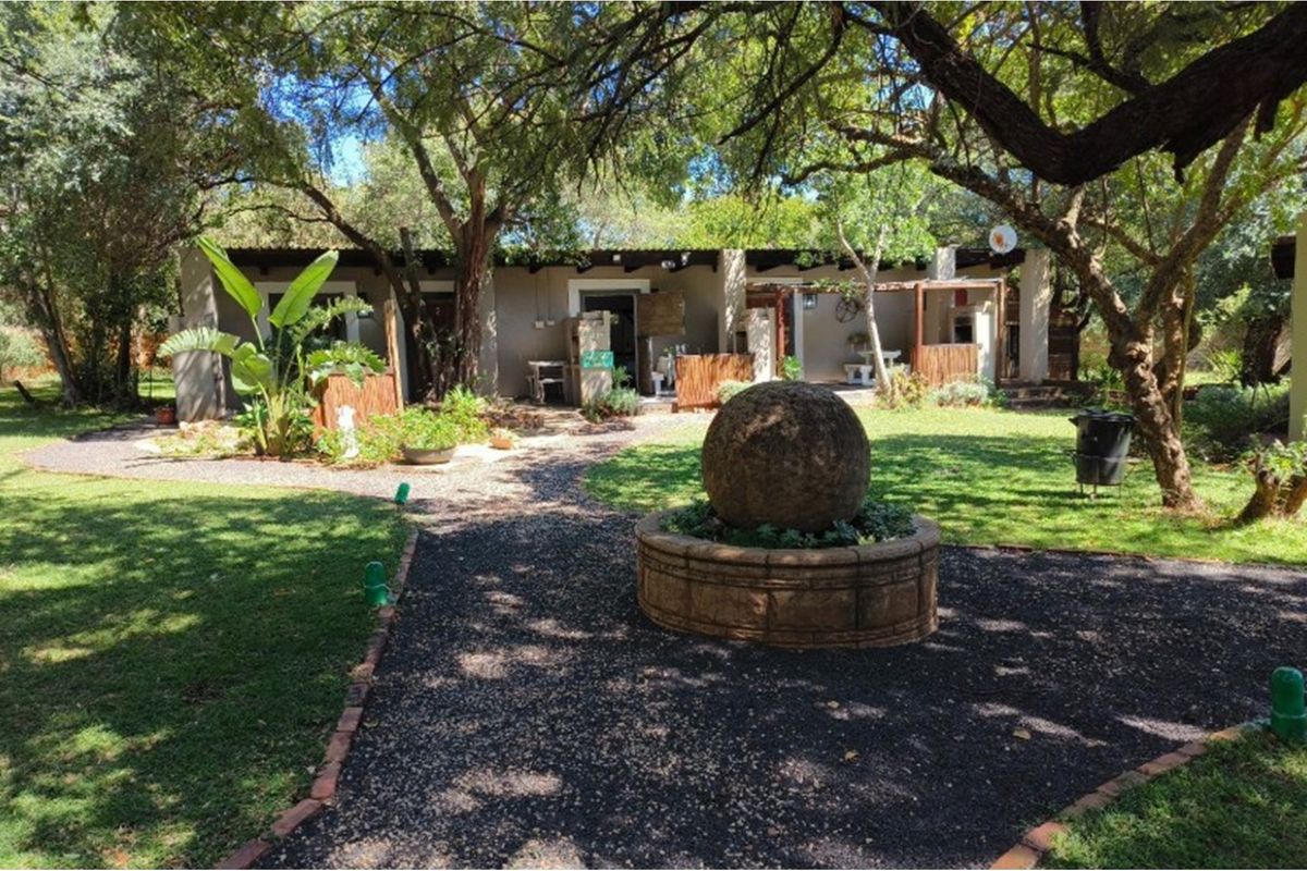 A tranquil rear garden area featuring a stone water feature, gravel pathways, mature trees, and a covered patio with a built-in braai.