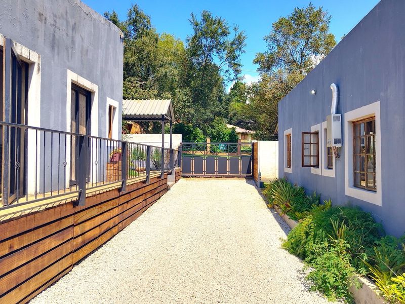 A gravel driveway leading to a secure gated entrance, flanked by two modern grey buildings with timber decking and landscaped garden beds.