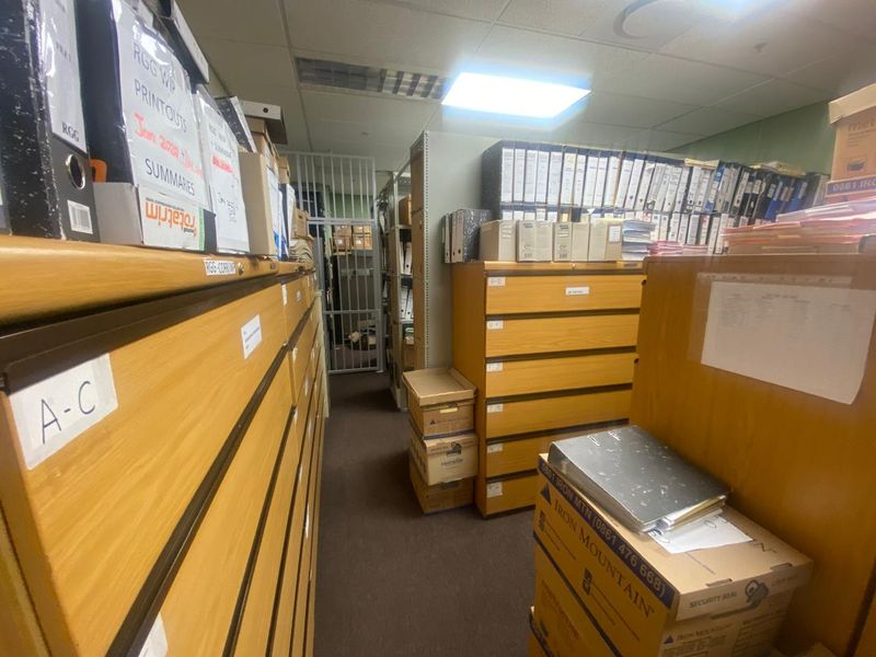 A functional office utility or storage room featuring built-in filing cabinets, carpeted flooring, and a secure metal gate entrance.