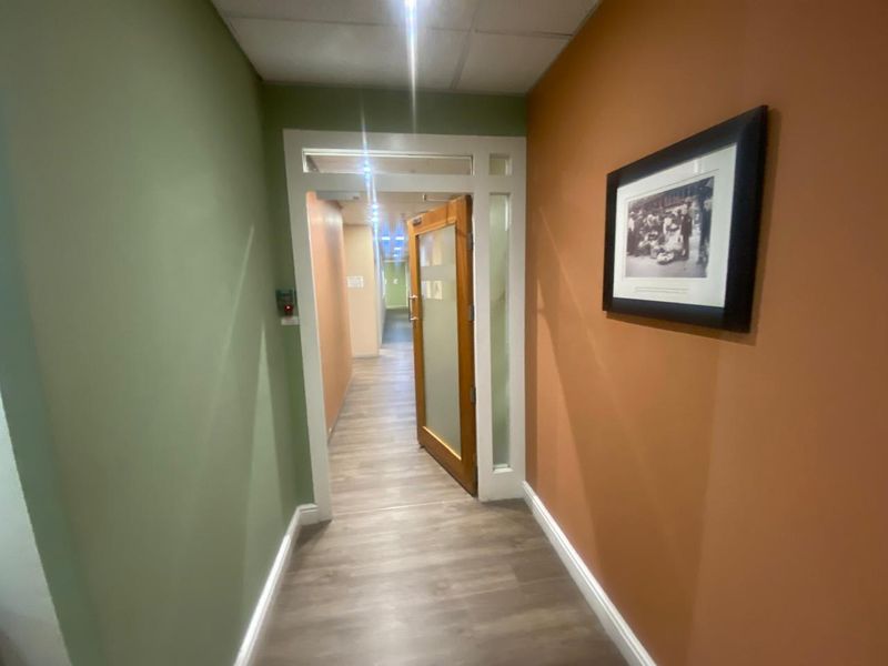 A well-maintained office hallway featuring laminate flooring, recessed ceiling lights, and a glass-paneled timber door leading to further office space.