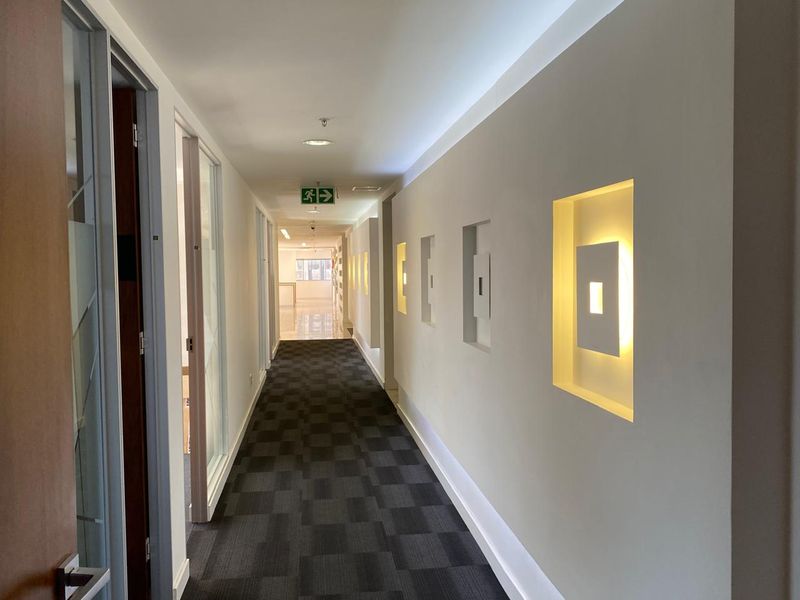 A modern office hallway featuring recessed wall lighting, carpet tile flooring, and glass partition doors leading to adjacent office spaces.