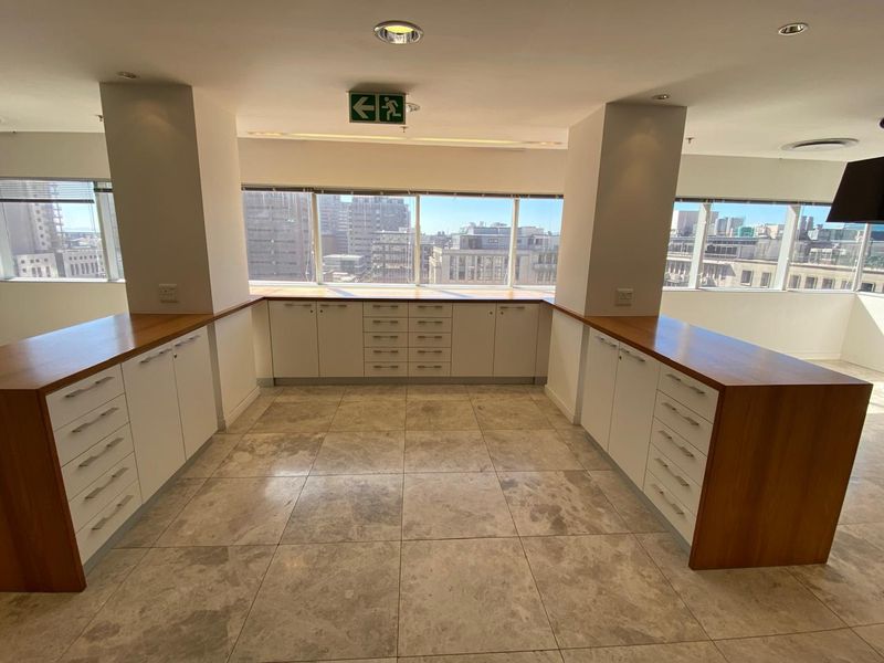 A bright, empty office space featuring built-in cabinetry with wooden countertops, stone tile flooring, and large windows offering city views.