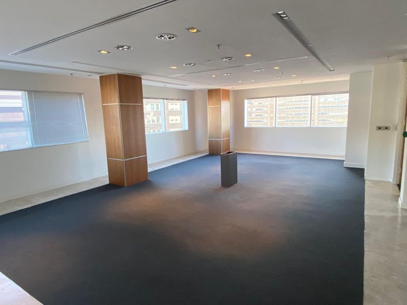 A spacious, open-plan office area featuring modern recessed lighting, wood-clad support columns, and large windows providing ample natural light.