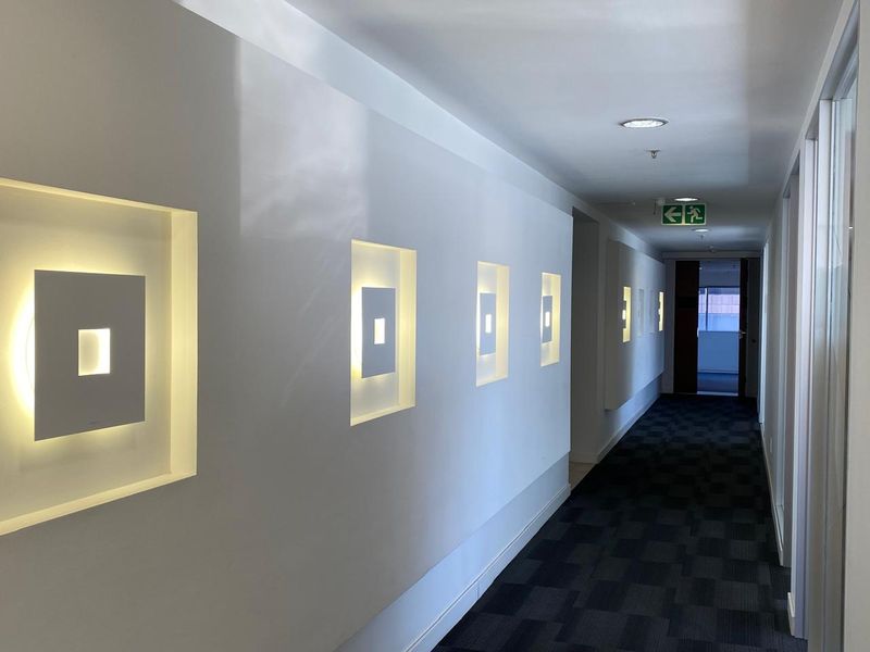 A modern office hallway featuring recessed wall lighting, dark patterned carpeting, and clear emergency exit signage.