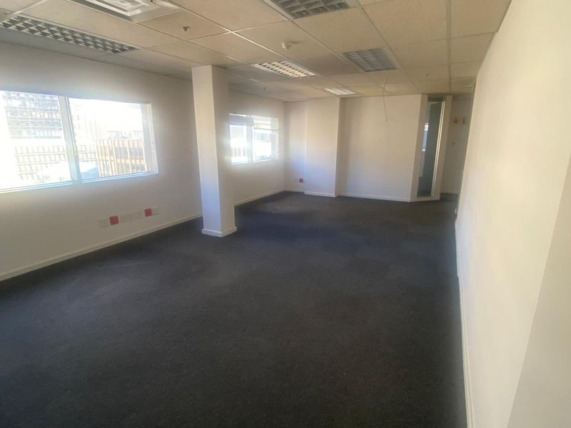 An empty, open-plan office space featuring neutral walls, carpeted flooring, and a suspended ceiling with recessed lighting.