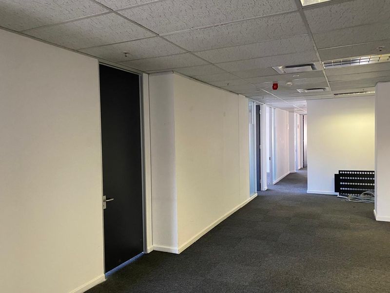 A spacious, empty office hallway featuring neutral walls, carpeted flooring, and a suspended ceiling with integrated lighting and climate control systems.
