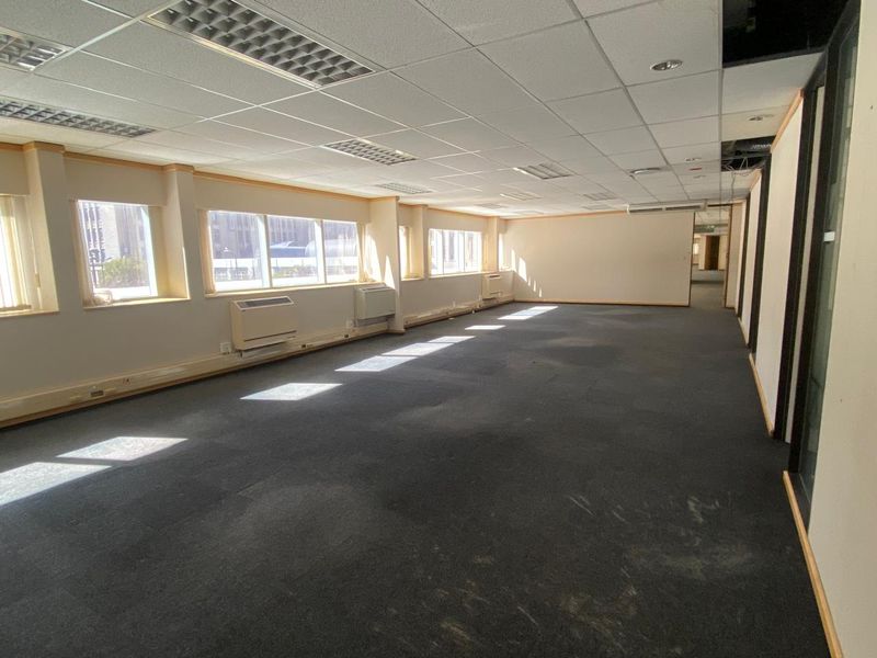 Spacious open-plan office area featuring suspended ceilings, recessed lighting, wall-mounted HVAC units, and large windows providing ample natural light.