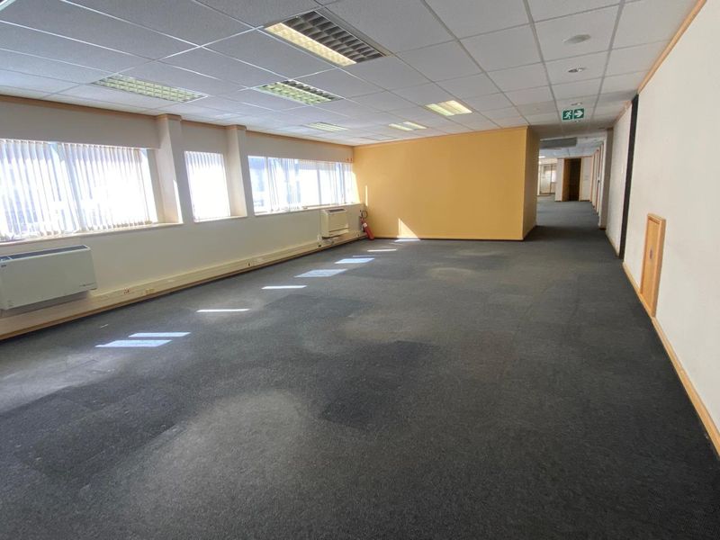Spacious, empty open-plan office area featuring suspended ceilings, recessed lighting, wall-mounted air conditioning units, and large windows with vertical blinds.