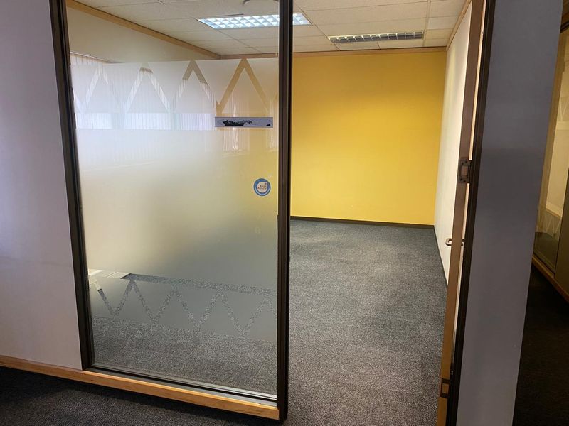 An empty office space featuring a glass partition with frosted detailing, carpeted flooring, and a suspended ceiling with recessed lighting.