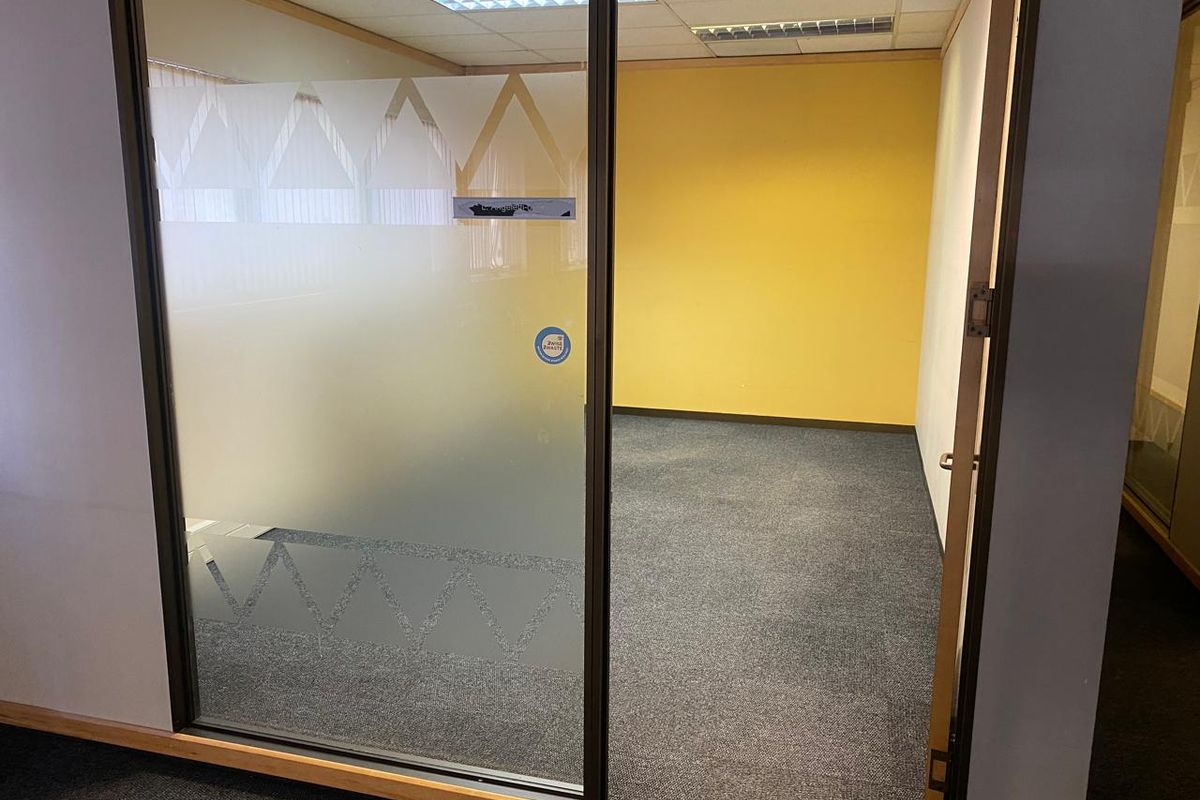 An empty office space featuring a glass partition with frosted detailing, carpeted flooring, and a suspended ceiling with recessed lighting.