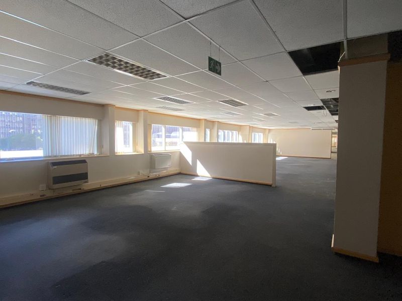 Spacious open-plan office area featuring suspended ceilings, recessed lighting, wall-mounted HVAC units, and large windows with vertical blinds.
