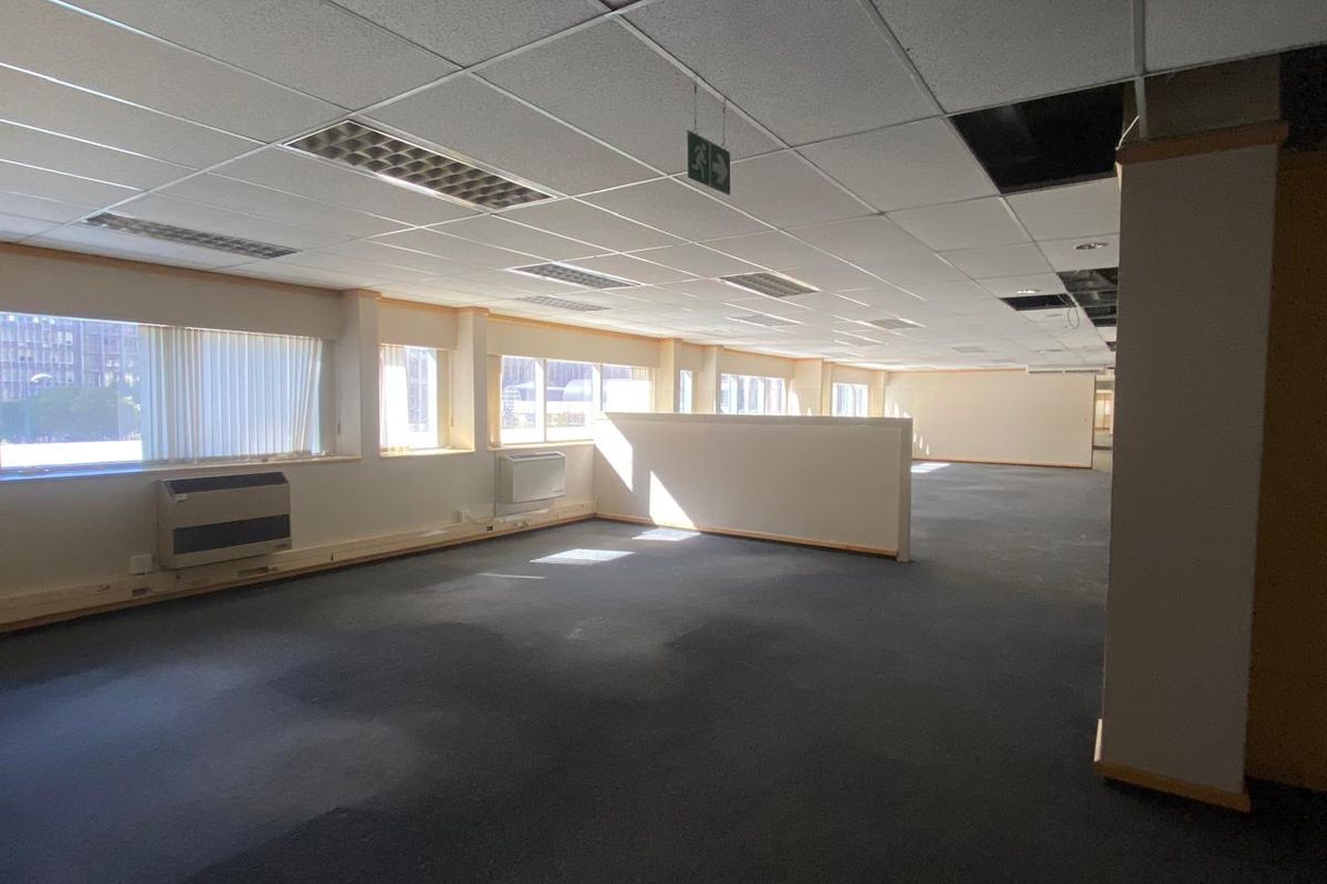 Spacious open-plan office area featuring suspended ceilings, recessed lighting, wall-mounted HVAC units, and large windows with vertical blinds.