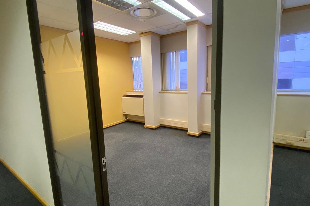 A vacant office space featuring carpeted flooring, suspended ceiling with integrated lighting, and a glass partition door.