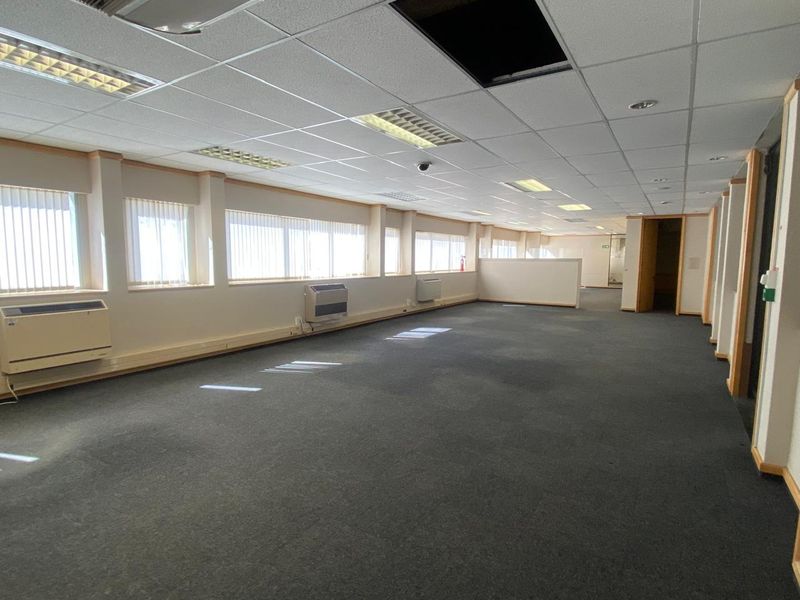Spacious open-plan office area featuring suspended ceilings, recessed lighting, wall-mounted HVAC units, and large windows with vertical blinds.