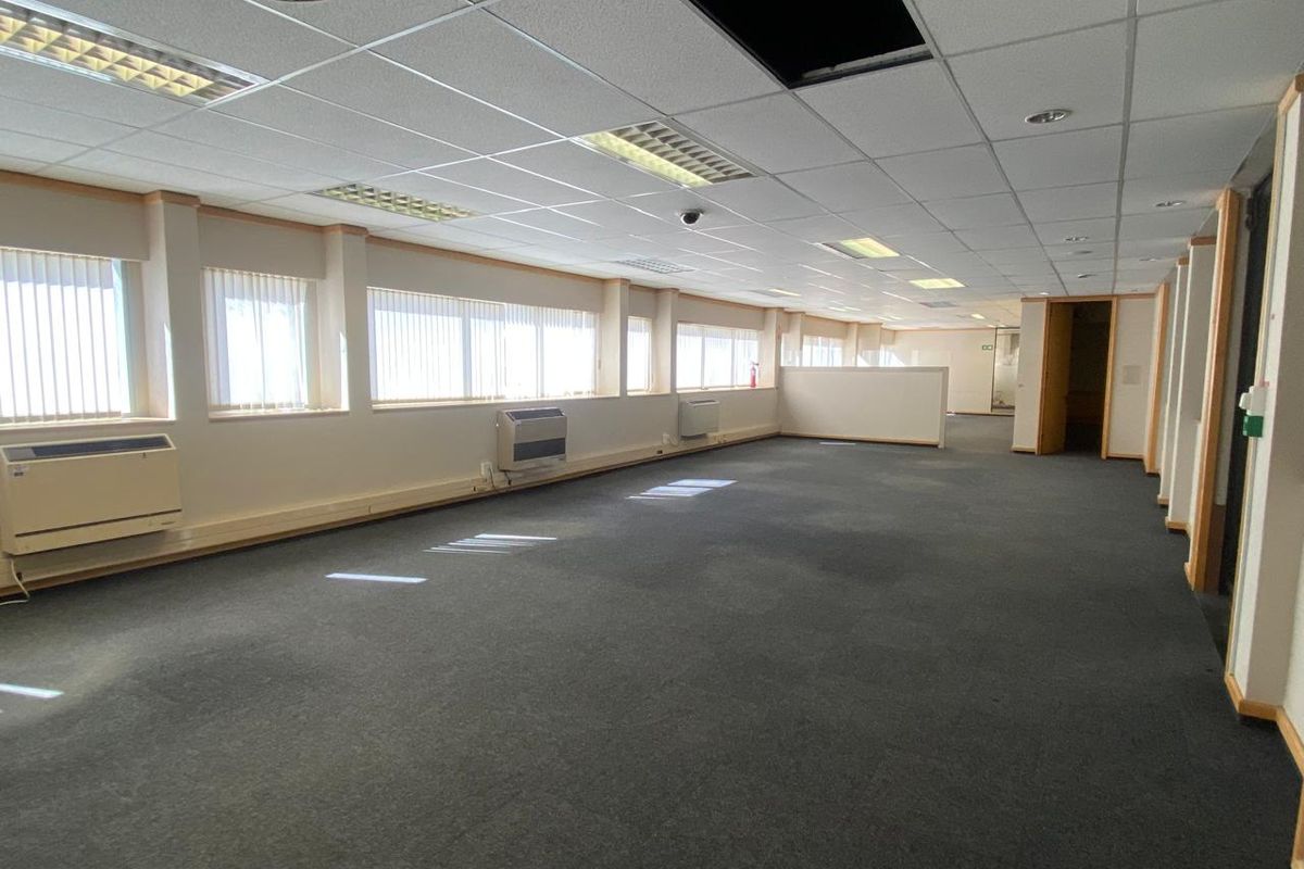Spacious open-plan office area featuring suspended ceilings, recessed lighting, wall-mounted HVAC units, and large windows with vertical blinds.