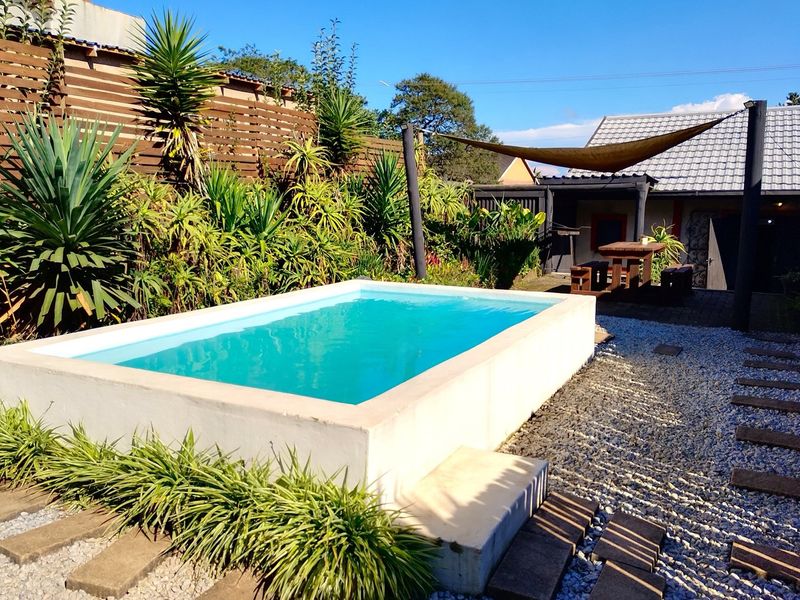 A well-maintained outdoor pool area featuring a raised concrete pool, gravel landscaping, and a shaded patio area with a wooden pergola.