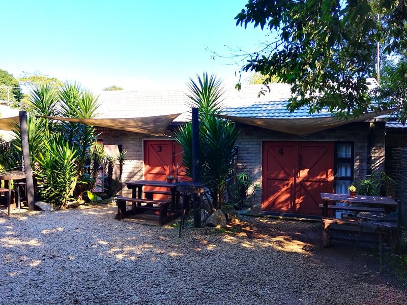 Exterior view of guest house units featuring brick facade, red double doors, and shaded outdoor seating areas with gravel ground cover.