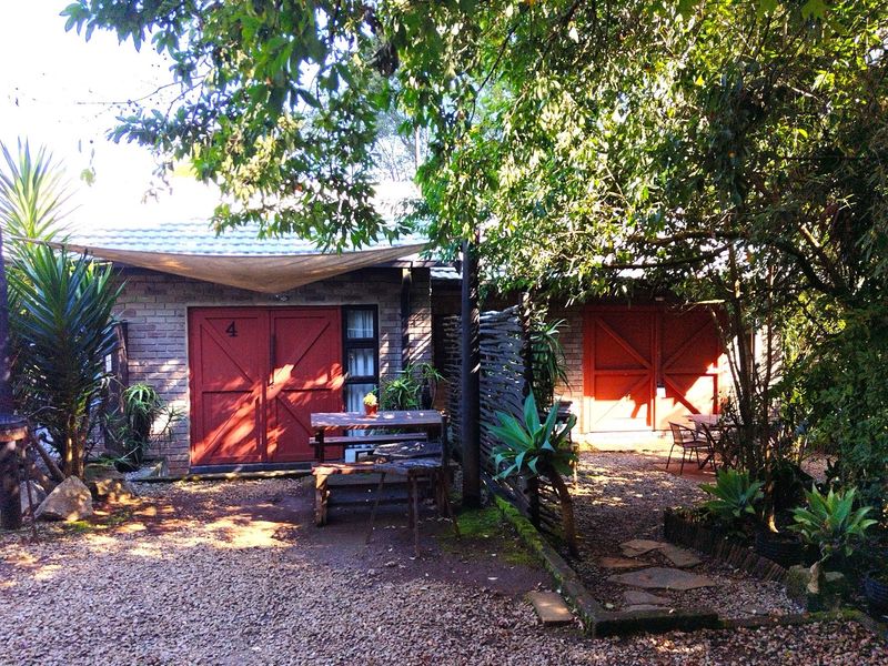 A rustic exterior view of guest house units featuring brick facades, red double doors, and a shaded gravel courtyard area.