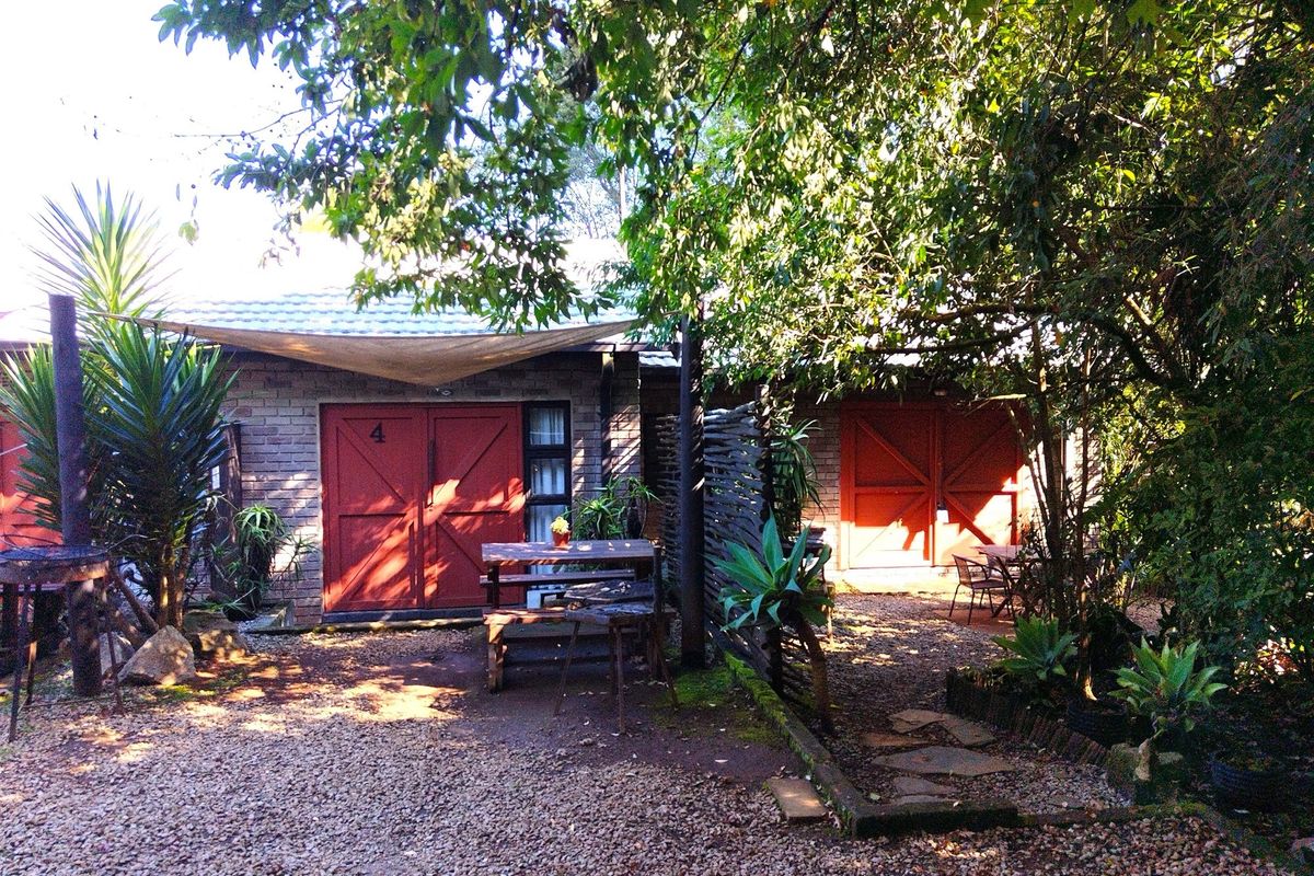 A rustic exterior view of guest house units featuring brick facades, red double doors, and a shaded gravel courtyard area.