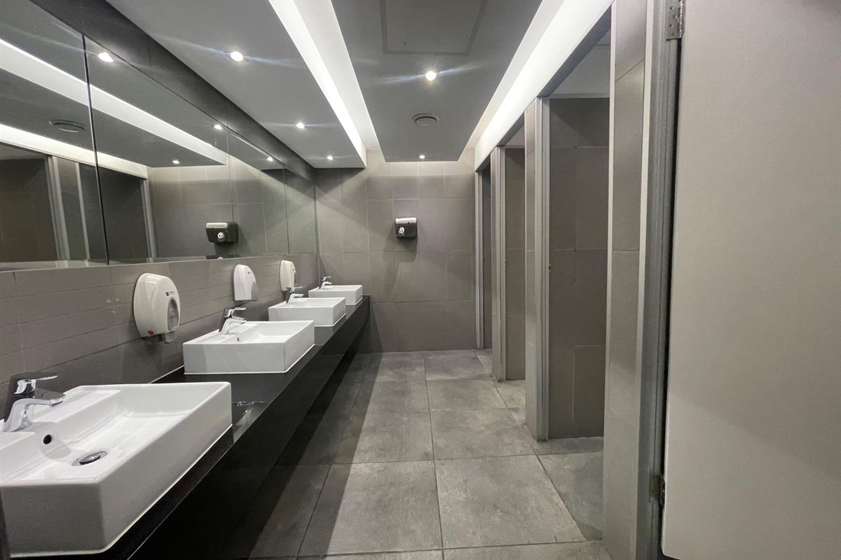 Modern commercial bathroom featuring multiple vessel sinks, large vanity mirror, recessed lighting, and private toilet cubicles with contemporary grey tiling.