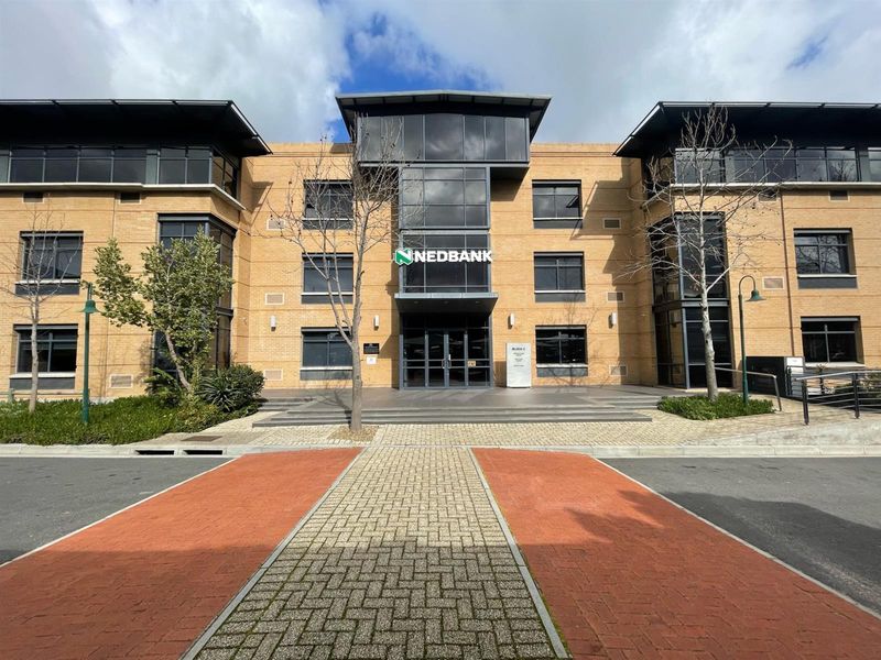 Front elevation of a modern multi-story office building featuring a brick facade, glass curtain wall entrance, and paved driveway access.