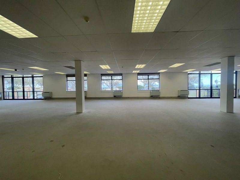 A spacious, open-plan office area featuring a suspended ceiling, recessed lighting, large windows with natural light, and integrated HVAC units.