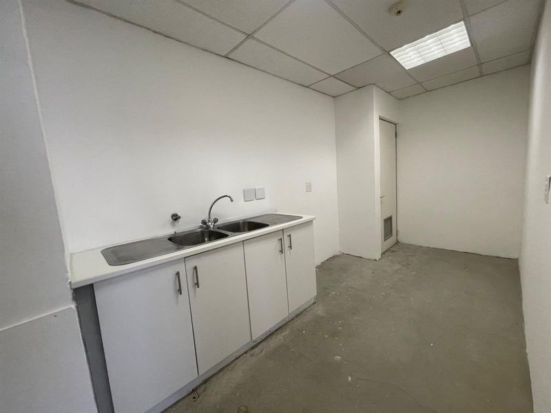 A small office kitchenette featuring white cabinetry, a stainless steel sink, and a drop ceiling, currently awaiting final flooring installation.