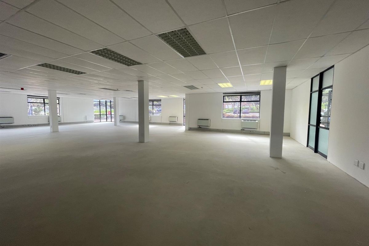A spacious, open-plan office area featuring a suspended ceiling, recessed lighting, structural columns, and large windows providing ample natural light.