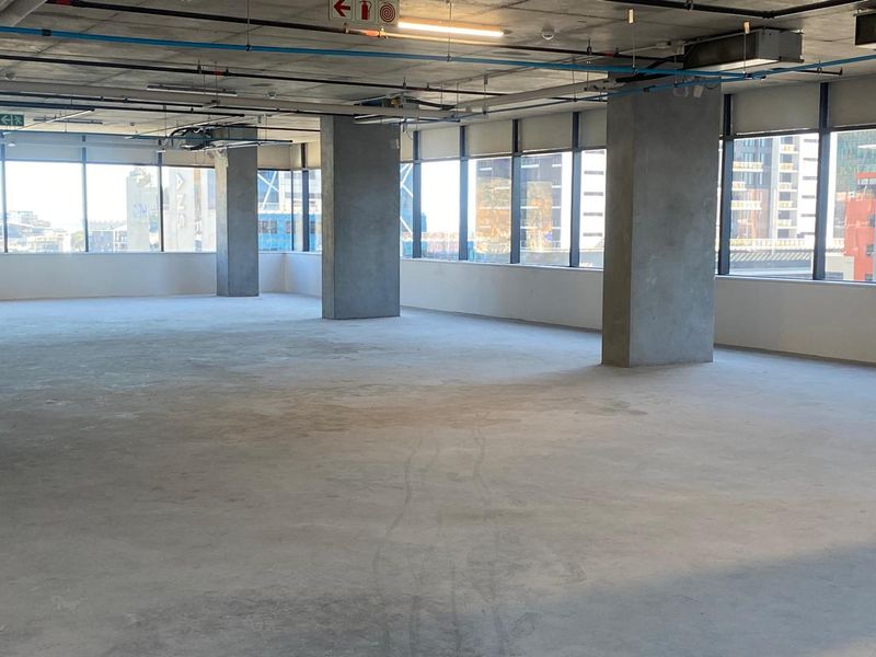 An expansive, open-plan office space featuring floor-to-ceiling windows, exposed concrete structural columns, and an industrial-style ceiling with visible services.