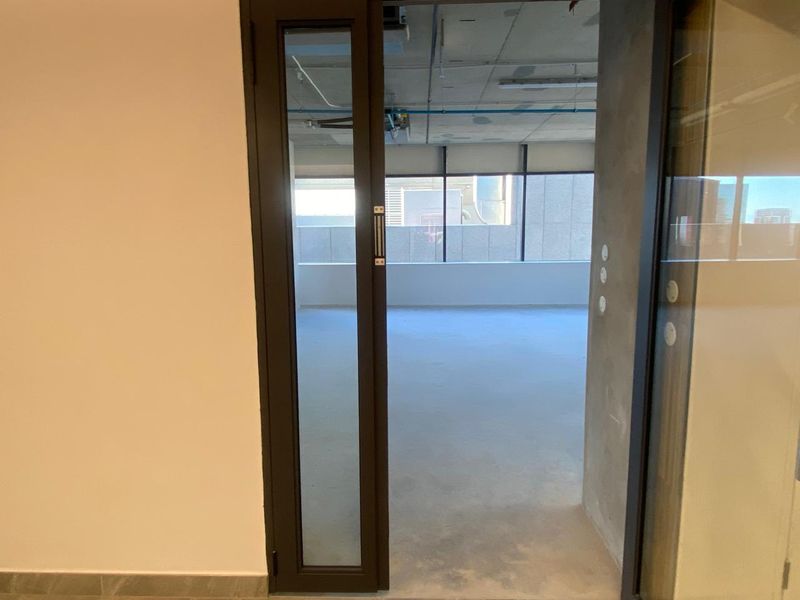 A view into an empty, open-plan office space featuring large windows, exposed concrete ceiling, and modern dark-framed glass partitioning.