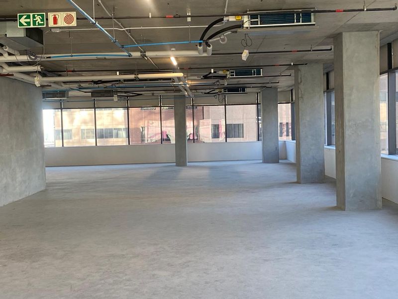 An open-plan office space featuring an industrial aesthetic with exposed ceiling infrastructure, concrete columns, and large windows providing ample natural light.
